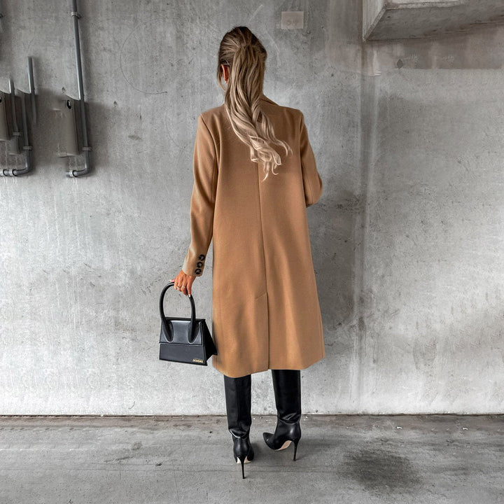Vrouw in stijlvolle camel winterjas, zwarte laarzen en handtas, staand tegen een betonnen muur. Wintermode, dameskleding, trendy outfit.