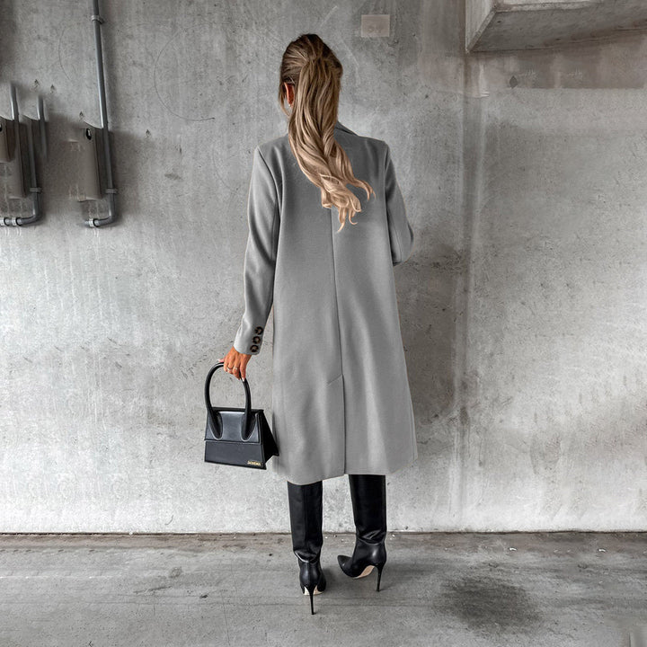 Vrouw in grijze winterjas, zwarte leren laarzen en handtas, staand tegen betonnen muur. Stijlvolle wintermode, minimalistisch design.