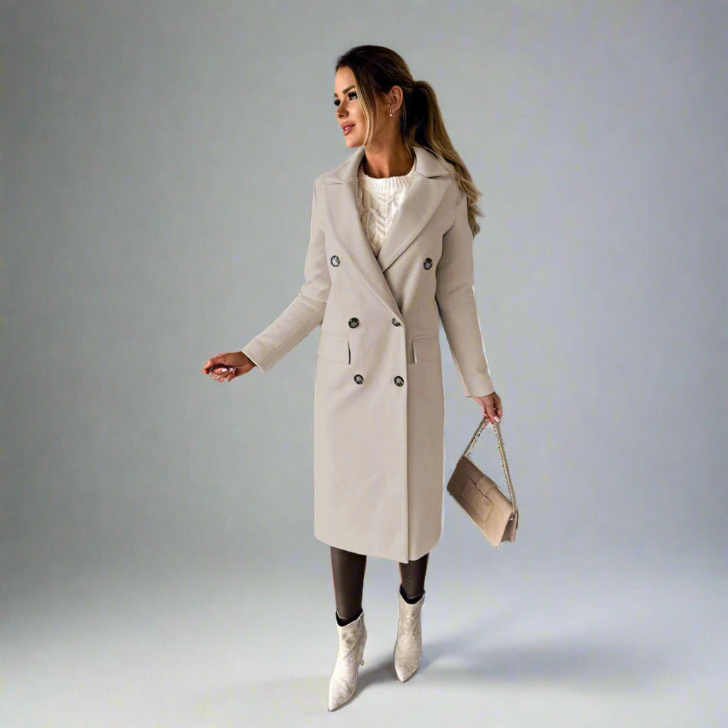 Vrouw in stijlvolle beige winterjas met dubbele rij knopen, witte trui en laarzen, poseert tegen een betonnen muur. Wintermode, dameskleding.