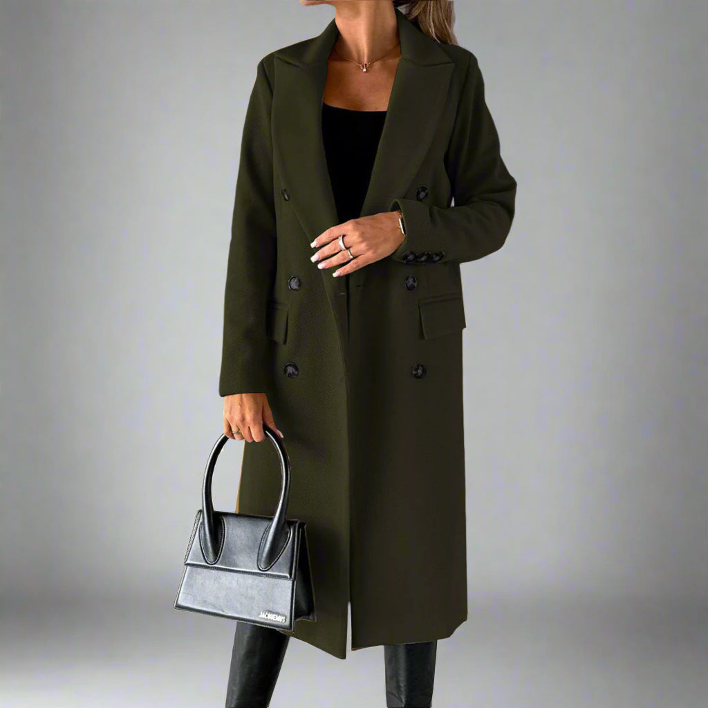 Vrouw in olijfgroene winterjas met dubbele knopen, zwarte handtas en zwarte laarzen tegen een grijze achtergrond. Stijlvolle damesmode.