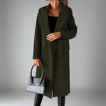 Vrouw in olijfgroene winterjas met dubbele knopen, zwarte handtas en zwarte laarzen tegen een grijze achtergrond. Stijlvolle damesmode.