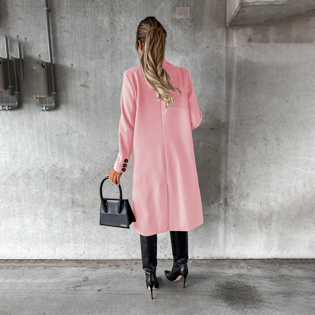 Vrouw in roze winterjas met zwarte laarzen en handtas, staand tegen een betonnen muur. Stijlvolle damesmode, winterkleding, trendy outfit.